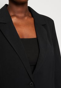 Blazer nero sartoriale con rever a lancia, chiusura con un solo bottone e tessuto liscio, indossato sopra un top nero aderente.