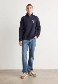 Hanorac navy cu fermoar și logo brodat, blugi gri-albastri și teniși bej cu accente maronii. Croială casual și manșete elasticizate.