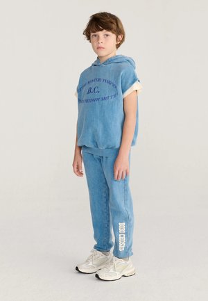 Un enfant se tient debout, portant un haut à capuche bleu à manches courtes et un pantalon assorti avec des baskets blanches sur un fond clair uni.
