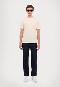 T-shirt beige chiaro a maniche corte, jeans dritti blu scuro e sneaker bianche. Il modello indossa occhiali da sole scuri, posato su uno sfondo neutro.