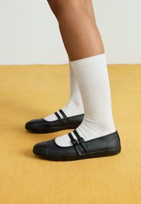 Sapatos de borracha pretos com duas correias e fivelas pretas, usados com meias brancas caneladas. Foco no design e textura do calçado.