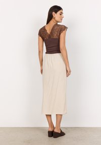 Braunes Spitzenoberteil mit kurzen Ärmeln, kombiniert mit einem beigefarbenen Midikleid. Das Oberteil hat eine taillierte Passform mit einem V-förmigen Rückenteil. Braune Blockabsatzschuhe.