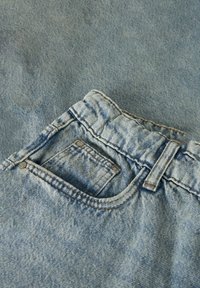 Svijetloplave traperice s vidljivim prednjim džepom, petljom za remen i detaljnim šavovima na teksturiranoj tkanini u pozadini.