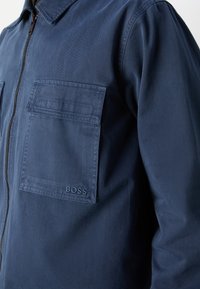 Gros plan d'une personne portant une veste zippée bleu marine avec une grande poche poitrine et le nom de la marque BOSS brodé dessus.