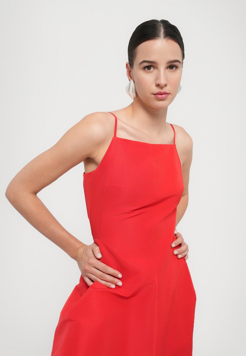 Une femme pose avec confiance dans une robe rouge à fines bretelles, les mains sur les hanches, mettant en valeur de grandes boucles d'oreilles stylées sur un fond neutre.