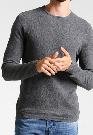 Mężczyzna w teksturowanym, szarym swetrze z długim rękawem i okrągłym dekoltem, stoi z rękami splecionymi na niebieskich dżinsach.