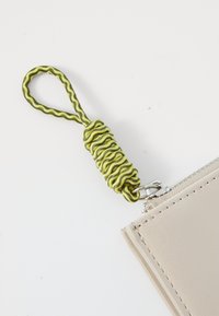 Pungă din piele bej cu închidere prin fermoar, având o curea spirale verzuie și neagră atașată prin hardware metalic.