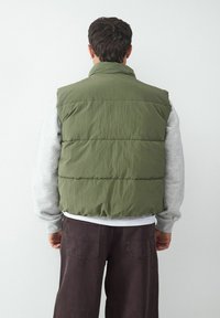 Olivgrüner Puffer-Weste mit hohem Kragen, gestepptem Design und leichtem Material. Getragen über einem grauen Langarmshirt. Dunkelbraune Hosen.