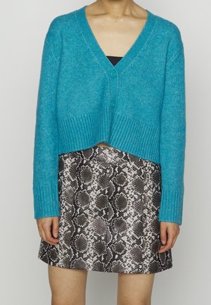 Femme portant un cardigan bleu court sur un haut noir, associé à une jupe courte à motif serpent noir et blanc.