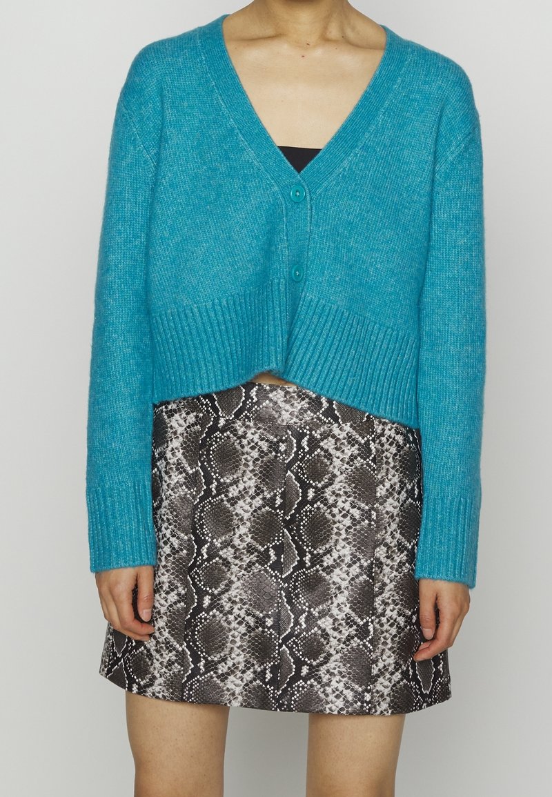 Femme portant un cardigan bleu court sur un haut noir, associé à une jupe courte à motif serpent noir et blanc.