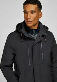 Veste isolante noire avec une fermeture éclair à l'avant et plusieurs poches à fermeture éclair. Dotée d'une capuche et de détails d'accentuation dans des couleurs contrastées.