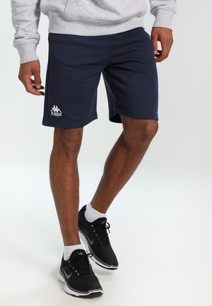 Mann trägt marineblaue Kappa-Shorts, grauen Pullover, weiße Socken und schwarze Nike-Sneaker und steht vor einem weißen Hintergrund.
