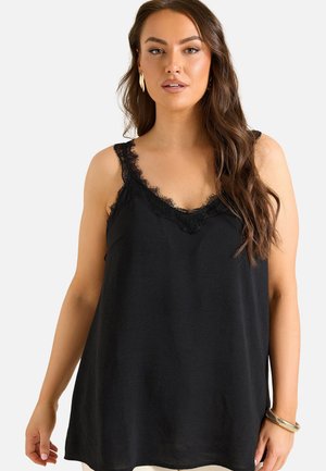 Mujer con cabello largo y castaño que lleva una blusa negra sin mangas con ribete de encaje y pendientes y pulsera dorados sobre un fondo claro y simple.