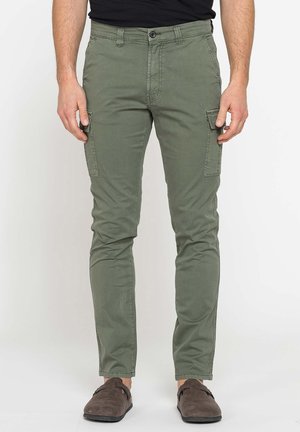 Uomo che indossa pantaloni cargo verde oliva slim fit con tasche laterali con bottoni e scarpe slip-on marrone scuro su sfondo bianco.