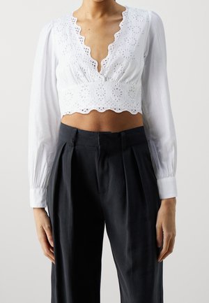 Femme portant un crop top blanc à manches longues avec des détails œillets et un pantalon noir plissé taille haute sur un fond uni.