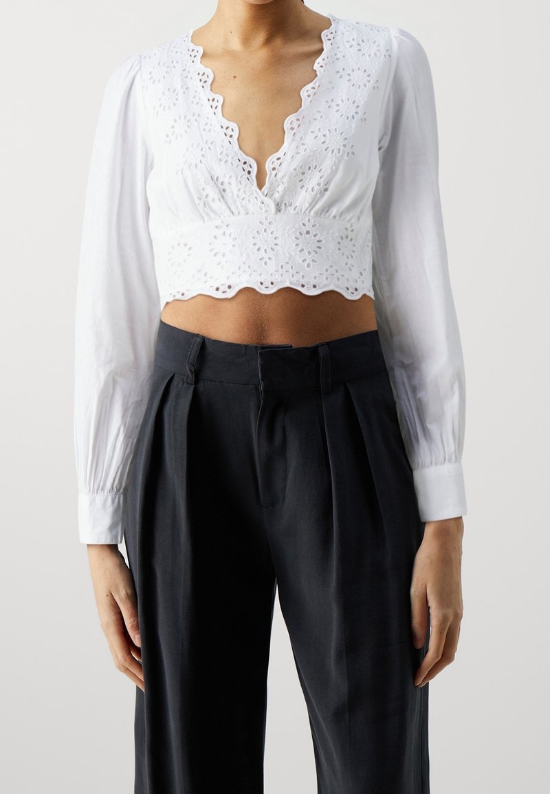 Femme portant un crop top blanc à manches longues avec des détails œillets et un pantalon noir plissé taille haute sur un fond uni.