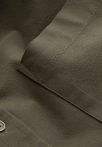 Olijfgroen stof, met een gestructureerd oppervlak, featuring naden en een metalen drukknoopsluiting. Close-up van het materiaal met minimale details.