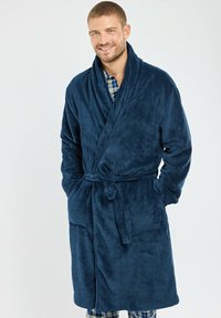Peignoir de bain en polaire bleu marine avec col châle, ceinture à nouer, deux poches avant et texture douce. Porté par-dessus une chemise à carreaux et un pantalon.