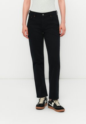 Persoon gekleed in zwarte rechte denim jeans en zwarte sneakers met witte veters en rubberen zolen, staand tegen een witte achtergrond.