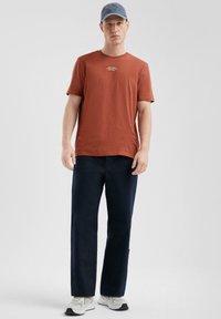 Roestkleurig T-shirt met korte mouwen en tekst. Donkerblauwe broeken, relaxte pasvorm. Lichtblauwe pet en witte sneakers met grijze accenten.