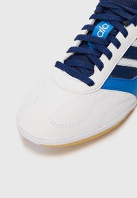 Bílé sportovní boty s modrými akcenty. Mají hladký syntetický svršek, ploché tkaničky a textura gumové podrážky pro lepší přilnavost.