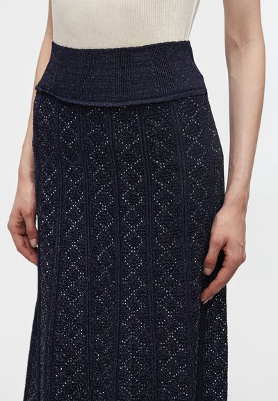 Personne portant un haut beige sans manches et une jupe tricotée bleu foncé avec des motifs texturés scintillants, les mains détendues sur les côtés.