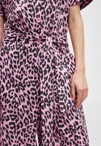 Rózsaszín leopárdmintás overall rövid ujjal, fekete részletekkel és megkötött derékkal. Nagy gombokkal és texturált anyaggal rendelkezik.