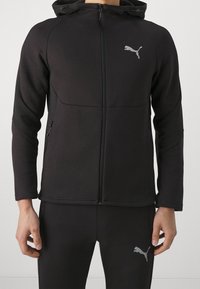 Chaqueta deportiva negra con cremallera frontal, capucha y bolsillos laterales. Presenta un diseño ajustado y un logo de Puma gris en el pecho, junto con pantalones a juego.