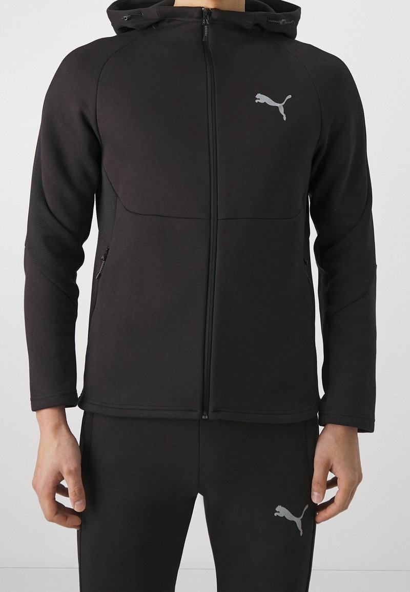 Giacca sportiva nera con zip frontale, cappuccio e tasche laterali. Presenta un design aderente e il logo Puma grigio sul petto, abbinata a pantaloni coordinati.