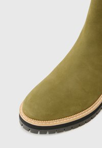 Grøn svinemateriale ankelstøvle med rund tå, kontrasterende beige syninger og en robust sort gummisål med et tekstureret mønster.