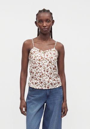 Femme aux cheveux tressés portant un haut blanc à fines bretelles fleuri et un jean bleu ample, debout devant un fond clair uni.