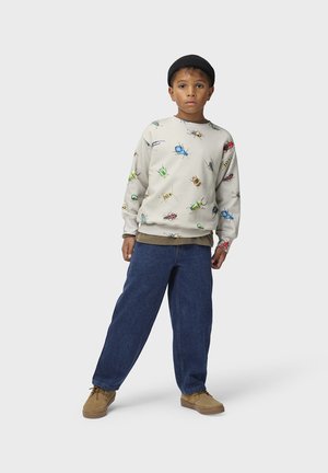 Dreng iført en beige sweatshirt med farverige insektmotiver, blå jeans, lyse sko og en sort hue, står foran en ensfarvet baggrund.