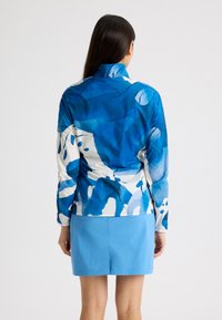 Långärmad jacka med ett blått och vitt abstrakt mönster, tillverkad av lätt tyg med hög krage. Matchad med en blå kjol.