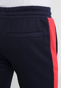 Marineblauwe joggingsbroek met een rode zijstripe, voorzien van een elastische tailleband en een klein achterzakje. Gladde stof met een aansluitende pasvorm.