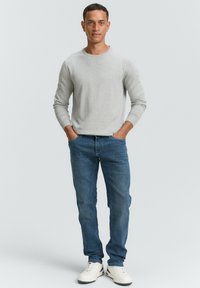 Lichtgrijze lange mouwen trui met een ronde hals, gecombineerd met medium blauwe spijkerbroek en witte sneakers, in een staande houding tegen een effen achtergrond.