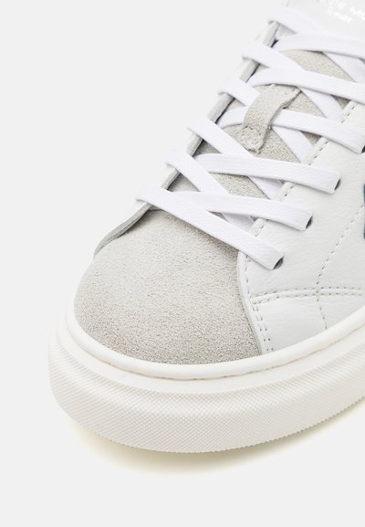 Zapatilla blanca con una puntera de gamuza gris, cordones blancos planos, parte superior de cuero texturizado y suela de goma con un dibujo en la banda de rodadura.