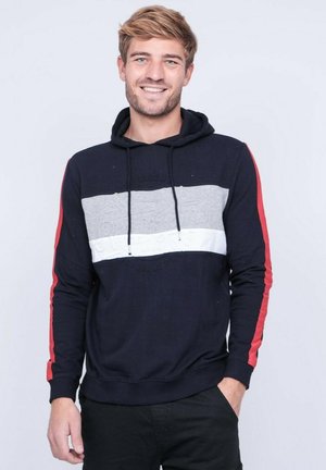Jeune homme souriant, portant un sweat à capuche noir avec des rayures grises et blanches et des rayures rouges sur les manches, debout avec une main dans la poche.