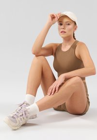 Haut court à côtes marron et shorts avec une casquette beige clair. Baskets légères avec des accents lavande et chaussettes blanches. Le fond est plain gris.