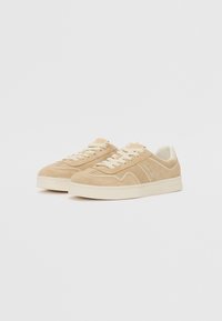 Sneakers in suede beige con lacci bianchi e suole in gomma. Il design presenta un motivo sottile sui lati e una forma della punta arrotondata.