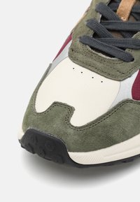 La sneaker présente un daim vert olive et des accents en cuir crème, une semelle en caoutchouc noir et des lacets gris. Le design comprend des détails perforés et une finition texturée.