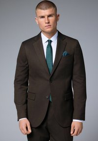 Costume marron avec une coupe ajustée, revers nets, deux poches avant, associé à une chemise blanche et une cravate turquoise, présentant un pochette.