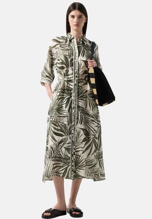 Kvinde iført lang kjole med knapper og grønne bladprint, der holder sort og beige stribet shoppertaske og har sorte sandaler på.