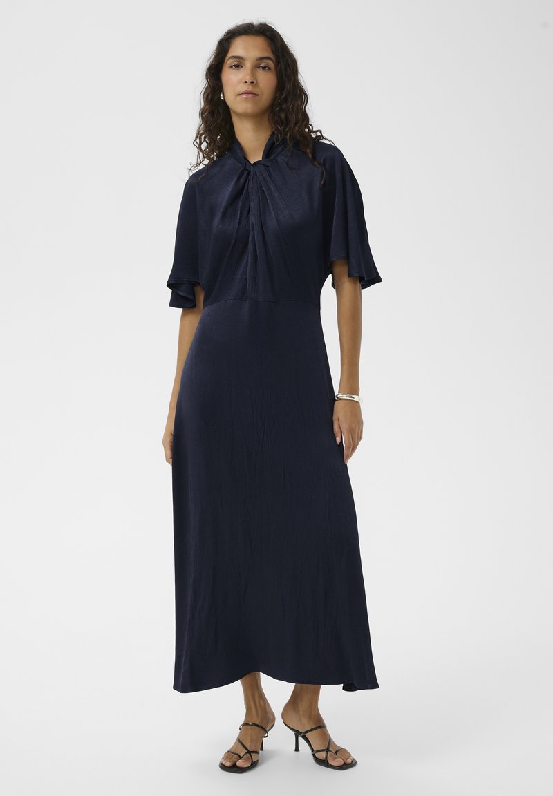 Maksi obleka mornarsko modre barve z mehko, teksturirano tkanino, z zavezovanjem okrog vratu in širokimi kratkimi rokavi. Oblečena s črnimi sandali s trakovi.