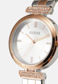 Montre ronde en argent avec des accents en or rose, cadran blanc texturé et embellissements en cristal sur la lunette et le bracelet. Marque : Guess.