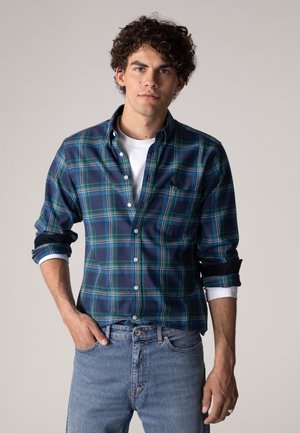 Ung mann med krøllete hår iført en blå og grønn rutete skjorte over en hvit t-skjorte og lyseblå jeans, stående med én hånd i lommen.