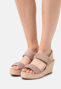 Beige kilehæle sandaler med ribbet stofremme og en vævet jutesål. Har åbne tæer og to justerbare ankelremme for støtte.