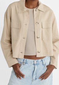 Cropped, light beige jacket in a plain fabric with a shirt collar, two front pockets, and button closure, paired with a ribbed top and denim shorts.