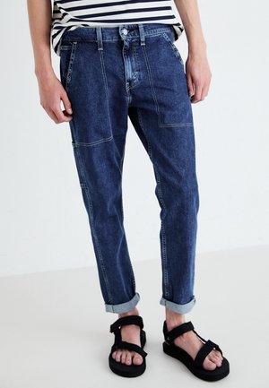 Jeans, dunkelblau, mit Kontrastnähten, umgeschlagenen Bündchen und einem entspannten Schnitt. Kombiniert mit schwarzen Sandalen mit Riemen.