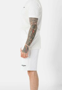 Maglietta bianca in cotone e pantaloni corti. La maglietta presenta un piccolo logo, mentre i pantaloni corti hanno il nome del marchio stampato. Tatuaggi colorati visibili sulle braccia.