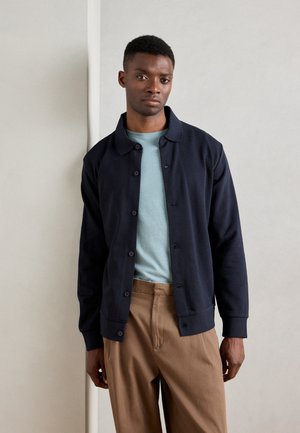 Navy button-up cardigan made of soft fabric, featuring a collared neckline and ribbed cuffs, paired with a light blue t-shirt and beige trousers.